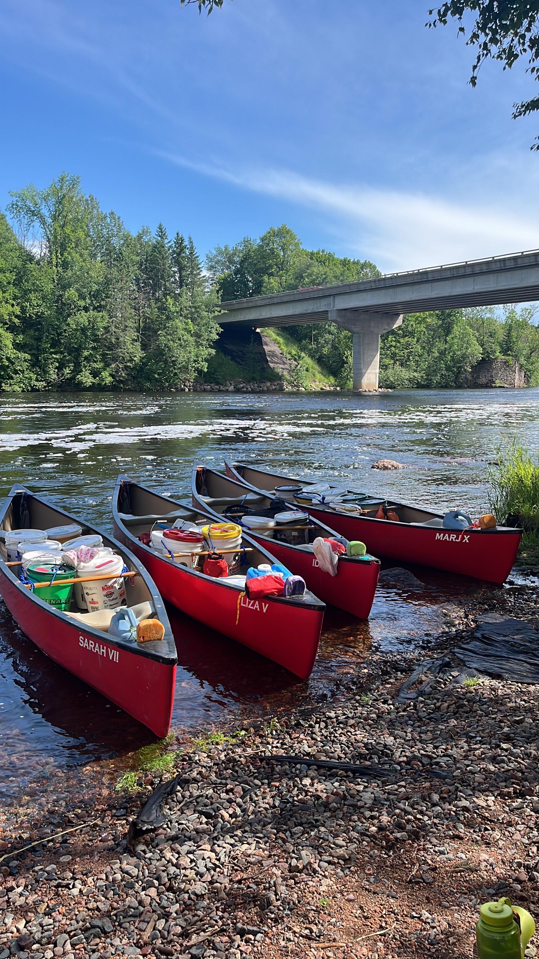 Four loaded red canoes pulled up on a stony shore beneath a Wisconsin bridge